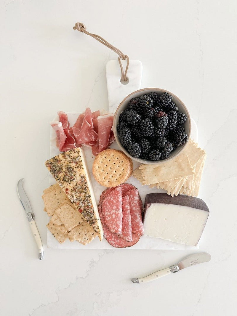 A gorgeous spread of food on top of a marble Caravan Home board featuring Laguiole cheese knives.
