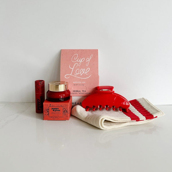 A collection of her favorite red gifts with from left to right this poppy and pout lip balm and scrub, cup of love tea for two, le bon shoppe her socks, and a nat and noor hair claw in cherry red.