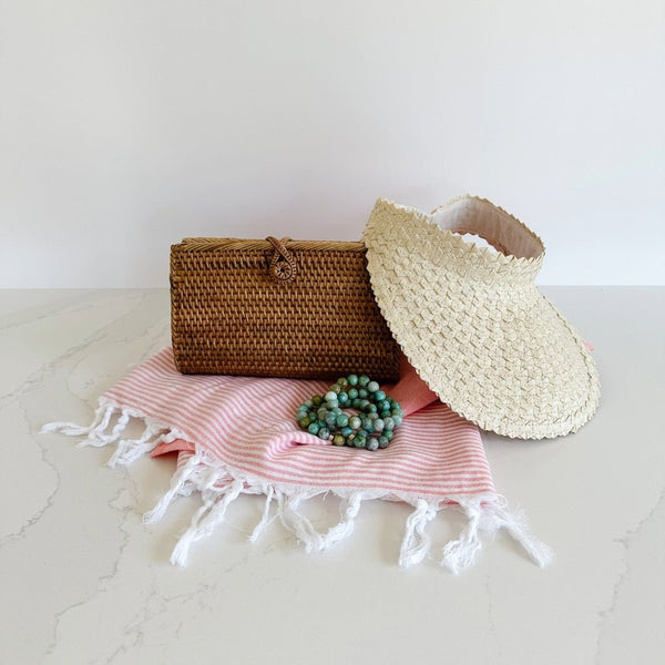 Turkish T Emma Breeze Towel with Etoiled Frog Leap beaded bracelets sitting on top of it. Behind the bracelets is a small brown rattan clutch from Bali Harvest and their Straw Sun Visor in a lighter hue is leaning off to the right. Great gift for her!