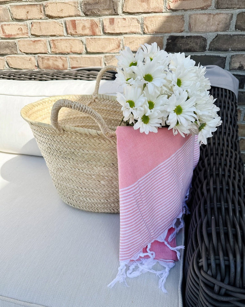 A neutral grass market basket from Bella Cucin with white flowers and a Emma Breeze coral colored striped Turkish towel from Turkish T sitting on a wicker chair with white cushions.