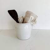 A beautiful crock from French Dry Goods holding a dark wooden spatula from Itza wood, a flax pot holder from Kate Kilmurray, and an oat checkered towel from state the label.