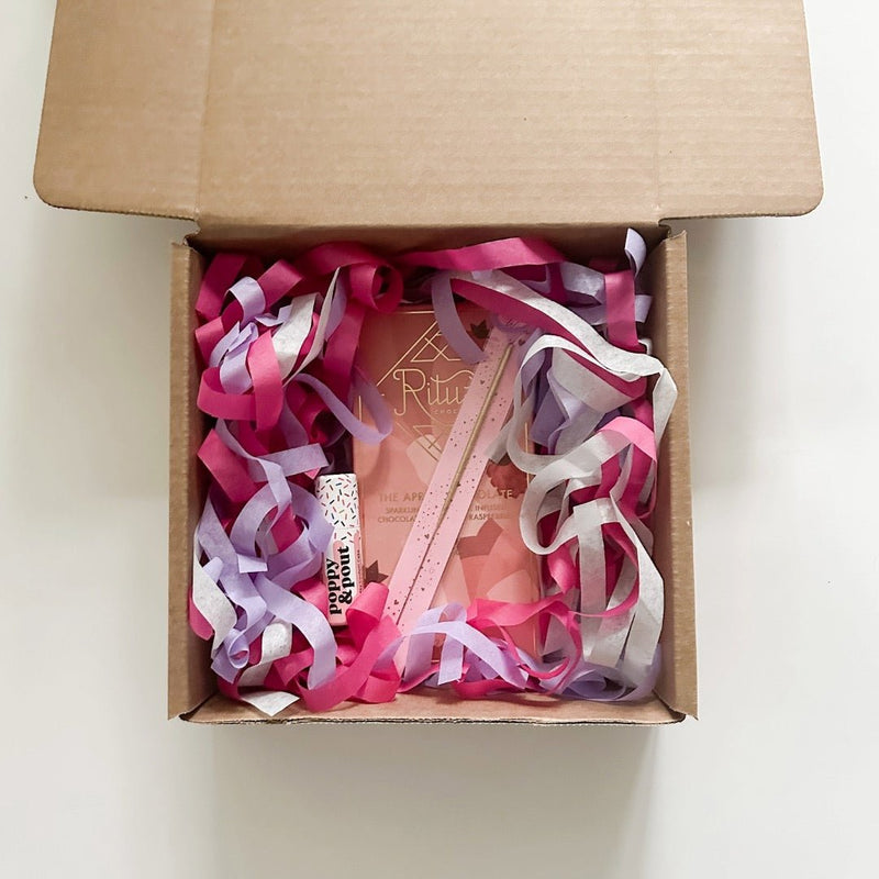 A fun Birthday gift box with hot pink, lavender and white strands of tissue paper and includes a Ritual Chocolate Aprés Chocolate Bar, Poppy and Pout birthday cake lip balm, and a single wish sparkler.