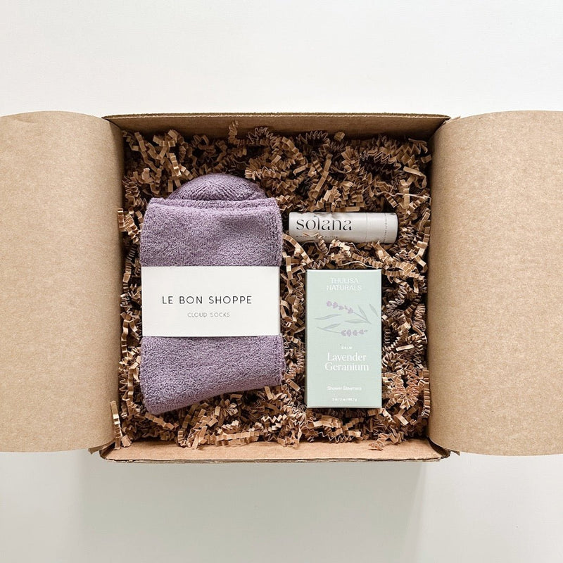 A self care gift box with le bon shoppe cloud socks in plum, Thulisa Naturals lavender geranium shower steamers, and Solana lip balm.